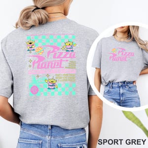 May include: Sport grey t-shirt featuring a Pizza Planet design. The design includes the text "Pizza Planet" in pink, along with cartoon alien characters and a checkered pattern in teal and pink. The shirt has a crew neck and short sleeves.