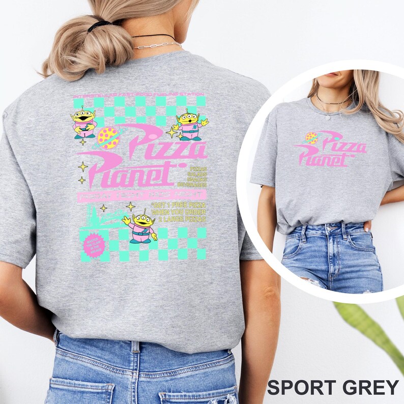 May include: Sport grey t-shirt featuring a Pizza Planet design. The design includes the text "Pizza Planet" in pink, along with cartoon alien characters and a checkered pattern in teal and pink. The shirt has a crew neck and short sleeves.
