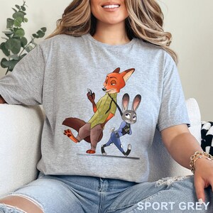 Pode incluir: Camiseta cinza esportiva com uma ilustra&ccedil;&atilde;o de desenho animado de Nick Wilde, uma raposa, e Judy Hopps, uma coelha, de Zootopia. Nick est&aacute; com um telefone, e Judy est&aacute; andando ao lado dele. O texto "SPORT GREY" est&aacute; impresso na parte inferior.