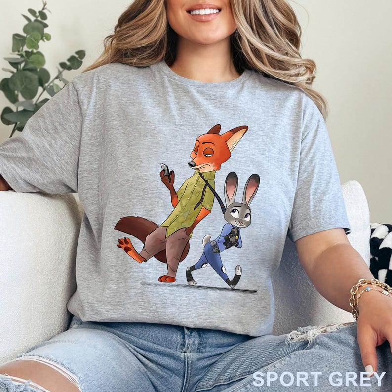Pode incluir: Camiseta cinza esportiva com uma ilustra&ccedil;&atilde;o de desenho animado de Nick Wilde, uma raposa, e Judy Hopps, uma coelha, de Zootopia. Nick est&aacute; com um telefone, e Judy est&aacute; andando ao lado dele. O texto "SPORT GREY" est&aacute; impresso na parte inferior.