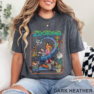 Pode incluir: T-shirt cinza escuro com um gr&aacute;fico colorido do filme de anima&ccedil;&atilde;o Zootopia. O design inclui o t&iacute;tulo do filme e v&aacute;rios personagens, incluindo uma raposa, um coelho e uma pregui&ccedil;a. O texto "DARK HEATHER" est&aacute; impresso na parte inferior.