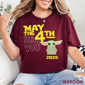 May include: A maroon t-shirt with a yellow graphic that says "May the 4th Be With You" and features a green cartoon character. The year 2025 is printed below the character.