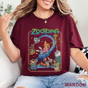 Pode incluir: Camiseta bord&ocirc; com a palavra "Zootopia" em verde e um desenho animado de personagens do filme. O desenho inclui uma raposa, um coelho e outros animais. A palavra "MAROON" est&aacute; impressa no canto inferior direito.
