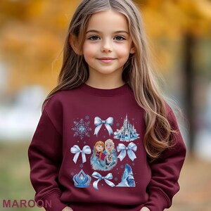 Puede incluir: Una sudadera granate con un dise&ntilde;o de los personajes Elsa y Anna de la pel&iacute;cula Frozen, copos de nieve, lazos y un castillo. La palabra "MAROON" est&aacute; impresa en la parte inferior.