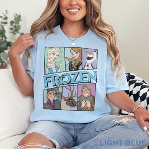 May include: Light blue t-shirt featuring a graphic of characters from the movie "Frozen." The design includes Elsa, Anna, Olaf, Kristoff, Sven, and a man in a hat. The word "FROZEN" is printed in blue. The shirt is labeled "LIGHT BLUE" at the bottom.