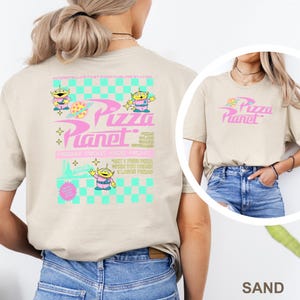 May include: Sand-colored t-shirt featuring the text "Pizza Planet" in pink, with a graphic of a pizza slice. The back of the shirt has a larger design with alien characters and the words "Pizza Planet" and "Friday Night Kids Night."