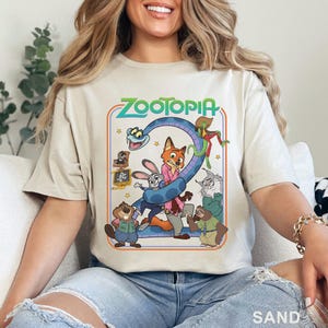 Pode incluir: Camiseta cor de areia com um gr&aacute;fico colorido do filme de anima&ccedil;&atilde;o "Zootopia". O design inclui o t&iacute;tulo do filme em verde, juntamente com v&aacute;rios personagens animais como uma raposa, um coelho e uma pregui&ccedil;a. A camisa &eacute; feita de material macio.