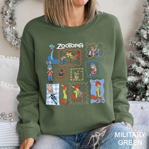 May include: Military green sweatshirt featuring a colorful graphic of characters from the animated film "Zootopia." The design includes the film's title and various character illustrations within decorative frames. The sweatshirt is a crew neck style.