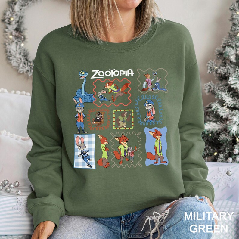 May include: Military green sweatshirt featuring a colorful graphic of characters from the animated film "Zootopia." The design includes the film's title and various character illustrations within decorative frames. The sweatshirt is a crew neck style.