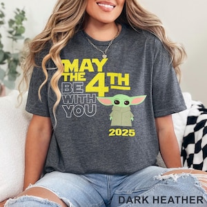 May include: Dark heather grey t-shirt with a yellow graphic that says "May the 4th Be With You" and features a green cartoon character. The year 2025 is printed below the character.