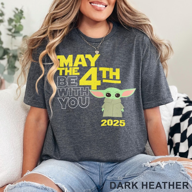 May include: Dark heather grey t-shirt with a yellow graphic that says "May the 4th Be With You" and features a green cartoon character. The year 2025 is printed below the character.