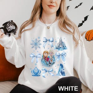 Puede incluir: Sudadera blanca con un dise&ntilde;o de acuarela de copos de nieve, lazos, un castillo y personajes. La palabra "WHITE" est&aacute; impresa en la parte inferior. Es una prenda c&oacute;moda de manga larga.