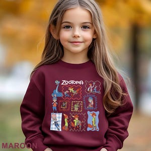 May include: A maroon sweatshirt featuring the word "Zootopia" and various character illustrations in colorful frames. The sweatshirt is worn by a young girl with long brown hair, set against a blurred autumn background.