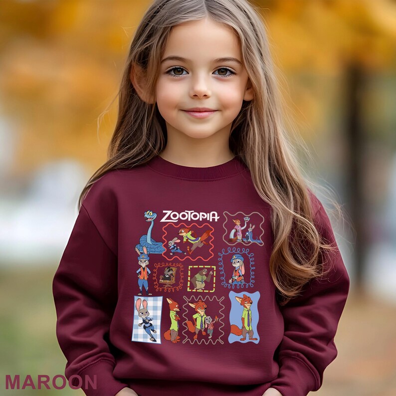 May include: A maroon sweatshirt featuring the word "Zootopia" and various character illustrations in colorful frames. The sweatshirt is worn by a young girl with long brown hair, set against a blurred autumn background.