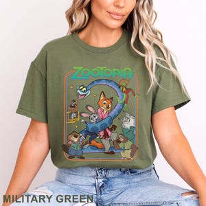 Pode incluir: Camiseta verde militar com um gr&aacute;fico colorido do filme de anima&ccedil;&atilde;o Zootopia. O design inclui o t&iacute;tulo do filme e v&aacute;rios personagens, como uma raposa, um coelho e uma pregui&ccedil;a. A camisa &eacute; um estilo casual e confort&aacute;vel.