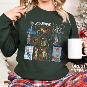 May include: Forest green sweatshirt featuring the word "ZOOTOPIA" and various character illustrations in colorful frames. The sweatshirt has a crew neck and long sleeves. A white mug is held in the hand.