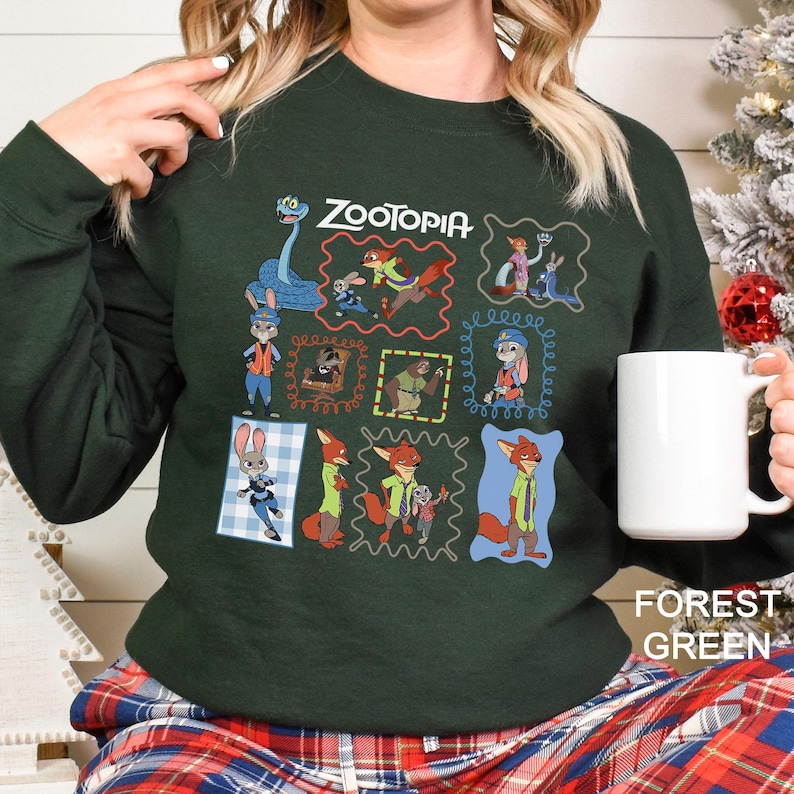 May include: Forest green sweatshirt featuring the word "ZOOTOPIA" and various character illustrations in colorful frames. The sweatshirt has a crew neck and long sleeves. A white mug is held in the hand.