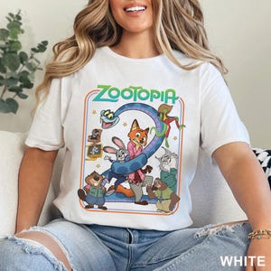 Pode incluir: Camiseta branca com um gr&aacute;fico colorido do filme de anima&ccedil;&atilde;o "Zootopia". O design inclui o t&iacute;tulo do filme e v&aacute;rios personagens de desenhos animados, incluindo uma raposa, um coelho e uma pregui&ccedil;a. A palavra "WHITE" &eacute; impressa na parte inferior.