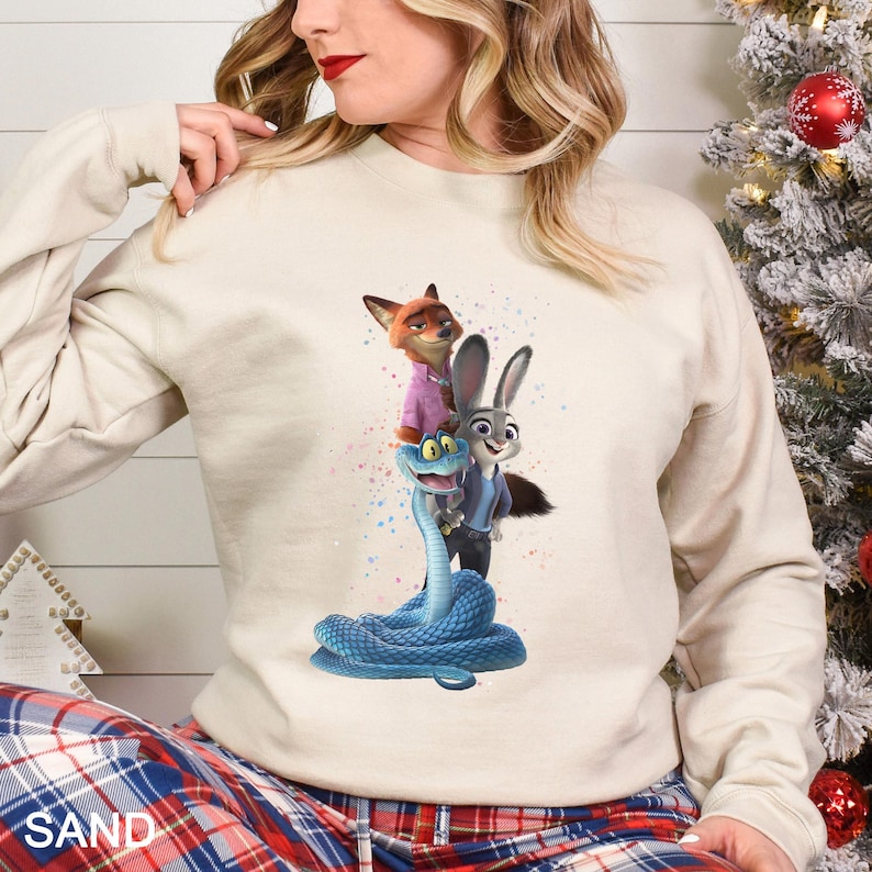 Peut inclure: Sweat-shirt couleur sable avec un graphisme des personnages du film d'animation Zootopie : Nick Wilde, Judy Hopps et un serpent bleu. Les personnages sont empil&eacute;s. Le mot "SAND" est imprim&eacute; en bas.