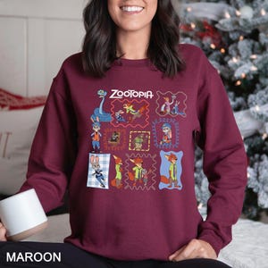 May include: Maroon sweatshirt featuring the word "Zootopia" above a grid of cartoon character images. The sweatshirt has a crew neck and long sleeves. The word "MAROON" is printed at the bottom of the image.