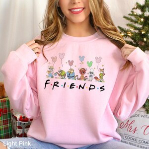 May include: Light pink sweatshirt featuring the word "FRIENDS" in a black font. Above the word, there is a cartoon illustration of several characters holding balloons. The balloons are in various colors, including pink, blue, and green.