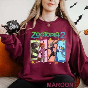 May include: A maroon sweatshirt with the text "Zootopia 2" and a graphic of characters from the animated film. The graphic includes a sloth, fox, rabbit, and snake. The word "MAROON" is printed at the bottom of the sweatshirt.