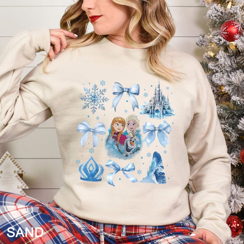 Puede incluir: Sudadera color arena con un dise&ntilde;o de la pel&iacute;cula Frozen, con la imagen de Anna, Elsa y Olaf, la palabra "SAND" en la parte inferior, la imagen de un castillo, lazos y copos de nieve azules.