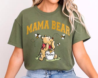 May include: An olive green short-sleeve t-shirt featuring the text MAMA BEAR in yellow arched letters. Below the text is a graphic of a yellow bear wearing a red shirt, holding a white pot labeled HUNNY, surrounded by three small flying bees.