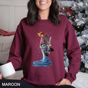 Peut inclure: Sweat-shirt bordeaux avec un motif de personnages anim&eacute;s : un renard, un lapin et un serpent bleu. Le renard porte une chemise rose, le lapin une veste bleue et le serpent est enroul&eacute;. Le mot "MAROON" est imprim&eacute; en bas.