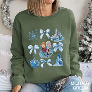Puede incluir: Sudadera verde militar con un dise&ntilde;o que presenta personajes y elementos de la pel&iacute;cula animada. El dise&ntilde;o incluye im&aacute;genes de Anna, Elsa, Olaf, copos de nieve, lazos, un castillo y una corona en tonos azules y blancos.