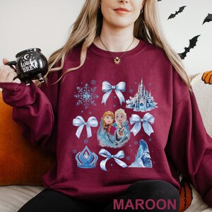 Puede incluir: Sudadera granate con un gr&aacute;fico de copos de nieve, lazos y un castillo. El gr&aacute;fico tambi&eacute;n incluye im&aacute;genes de dos princesas y un mu&ntilde;eco de nieve. La palabra "MAROON" est&aacute; impresa en la parte inferior de la sudadera.