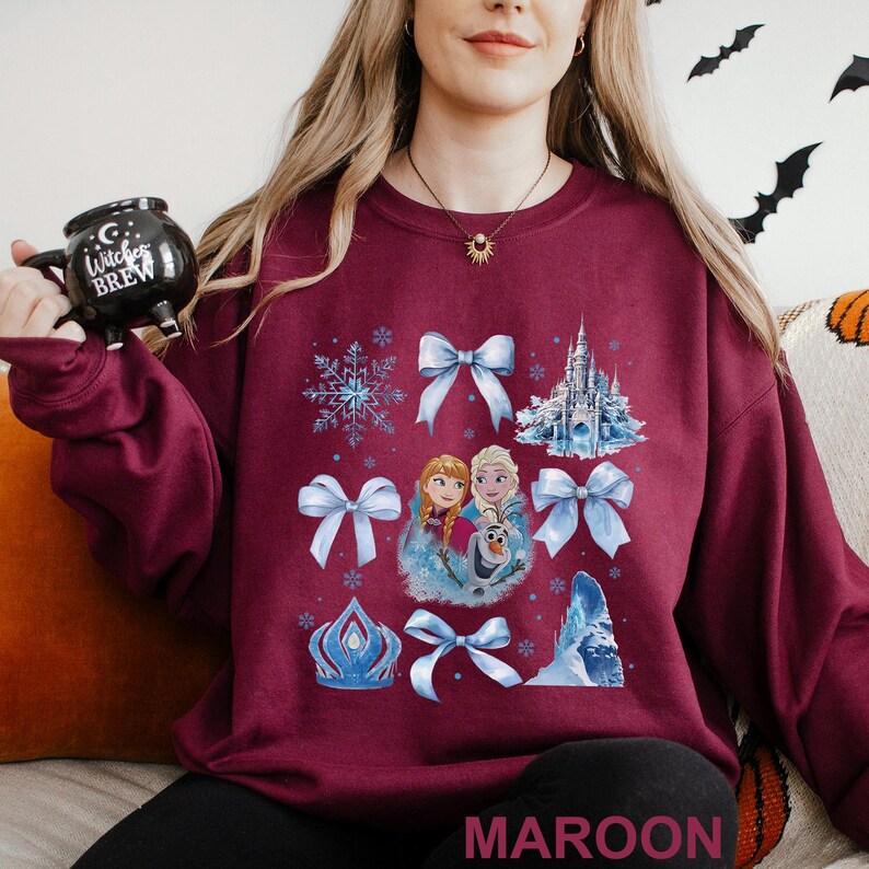Puede incluir: Sudadera granate con un gr&aacute;fico de copos de nieve, lazos y un castillo. El gr&aacute;fico tambi&eacute;n incluye im&aacute;genes de dos princesas y un mu&ntilde;eco de nieve. La palabra "MAROON" est&aacute; impresa en la parte inferior de la sudadera.