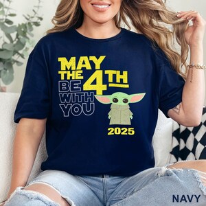May include: A navy blue t-shirt with a yellow graphic that says "May the 4th Be With You" and a cartoon image of a green alien character. The year 2025 is printed below the graphic.