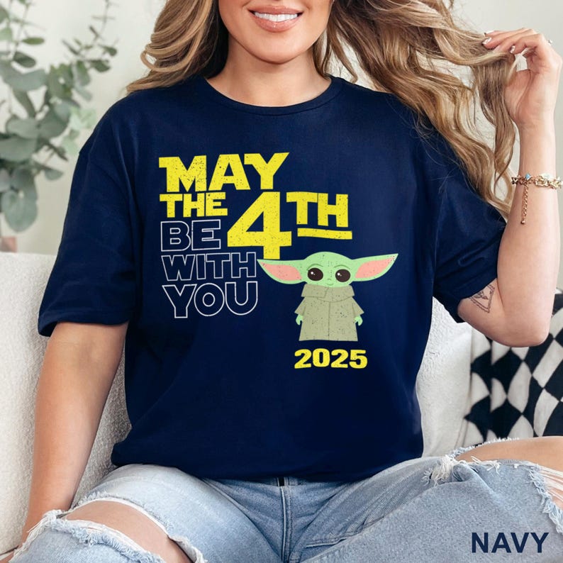 May include: A navy blue t-shirt with a yellow graphic that says "May the 4th Be With You" and a cartoon image of a green alien character. The year 2025 is printed below the graphic.