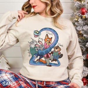 May include: A sand-colored sweatshirt featuring a cartoon graphic of characters from Zootopia. The design includes a blue snake forming the number 2, with characters like Judy Hopps, Nick Wilde, and others. The word "SAND" is printed at the bottom.