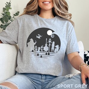 Pu&ograve; includere: T-shirt grigio sportivo con grafica in bianco e nero dei personaggi di The Nightmare Before Christmas. Il design include Jack Skellington, Sally e altre figure in una scena di cimitero. Il testo "SPORT GREY" &egrave; stampato in basso.