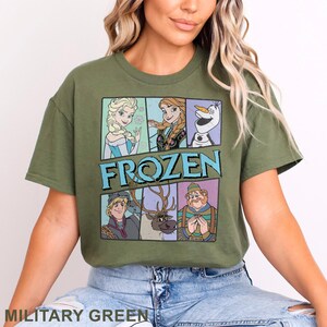 May include: Military green t-shirt featuring a graphic of characters from the movie "Frozen." The design includes Elsa, Anna, Olaf, Kristoff, Sven, and a shopkeeper. The word "FROZEN" is printed in large, blue letters. The shirt is a crew neck.