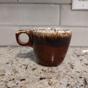 May include: A brown ceramic mug with a unique handle design. The mug features a gradient effect, transitioning from a light cream color at the rim to a deep brown at the base. The handle has a circular opening.