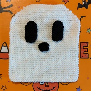 Könnte beinhalten: Weiße gehäkelte Geistermütze mit schwarzen Filzaugen und einer Nase. Die Mütze befindet sich auf einem orangefarbenen Hintergrund mit Halloween-Motiv.