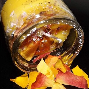 Könnte beinhalten: Eine gelbe Kerze mit getrockneten Rosenblüten und einem Docht in einem Glasgefäß. Das Gefäß ist umgekippt und die Blütenblätter fallen heraus.