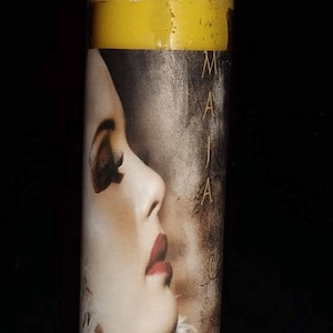May include: A yellow candle in a glass jar with a label featuring a woman's face and the word "MAJA" on it.