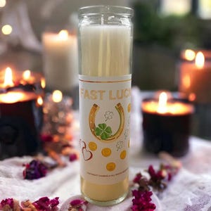 Könnte beinhalten: Eine weiße Kerze mit einem goldenen Etikett, auf dem "Fast Luck" steht und ein goldenes Hufeisen mit einem vierblättrigen Kleeblatt darin abgebildet ist. Das Etikett zeigt auch goldene Münzen und Herzen.