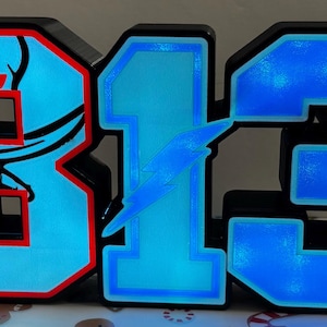 May include: Illuminated sign displaying the numbers "813" in a bold, blocky font. The "8" features a skull and football graphic, the "1" has a lightning bolt, and the "3" has a star. Each number glows with a bright blue light, outlined in black and red.