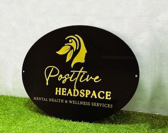 Puede incluir: Un letrero ovalado negro con letras doradas que dice "Positive Headspace Mental Health & Wellness Services". Una imagen dorada estilizada de una cabeza de persona con un diseño de hoja está por encima del texto.