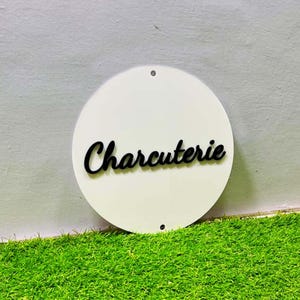 May include: A white, circular sign with the word "Charcuterie" in black script. The sign has two small holes at the top for hanging. The sign is set against a background of green artificial grass.