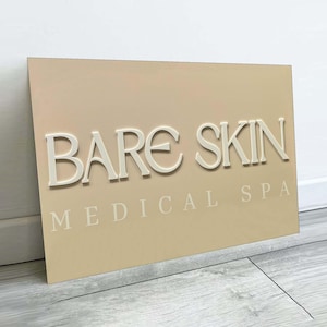 May include: A rectangular sign with the words "BARE SKIN MEDICAL SPA" in a raised, off-white font. The sign has a light beige background and is angled slightly, showing the corner. The sign is placed against a white wall and a light grey floor.