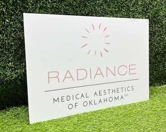 May include: White rectangular sign with the word "RADIANCE" in a dusty rose color, above the words "MEDICAL AESTHETICS OF OKLAHOMA" in black. A sunburst design is above the word "RADIANCE". The sign is on a green surface.