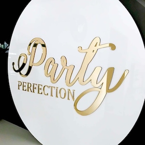 K&ouml;nnte beinhalten: Rundes wei&szlig;es Schild mit goldener Aufschrift "Party Perfection".