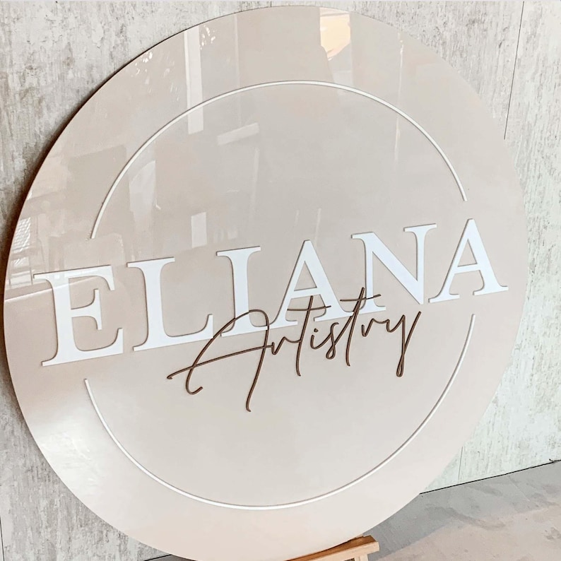 K&ouml;nnte beinhalten: Ein rundes, beiges Schild mit einem wei&szlig;en Kreis in der Mitte. Der Name "ELIANA" ist in wei&szlig;en Buchstaben &uuml;ber dem Kreis geschrieben, und das Wort "Artistry" ist in brauner Kursivschrift unter dem Namen geschrieben.