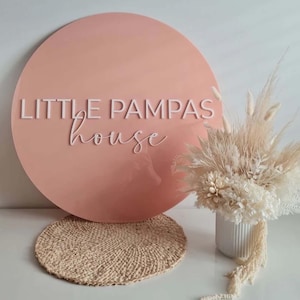 K&ouml;nnte beinhalten: Ein rundes rosa Schild mit dem Text "LITTLE PAMPAS house" in wei&szlig;en Buchstaben. Das Schild steht auf einer geflochtenen Matte. Eine wei&szlig;e Vase mit getrocknetem wei&szlig;en Pampasgras steht im Vordergrund.